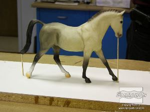 לחיצה להגדלת תמונה בעזרת דבק חם, מייצבים את הסוס על גבי לוח הבסיס. כדי למנוע לכידה של בועות אוויר בזמן היציקה, יוצרים תעלות אוורור במקומות אסטרטגיים על המודל. במקרה זה, קיסמי עץ מודבקים אל המודל בקצה הזנב ובראש.